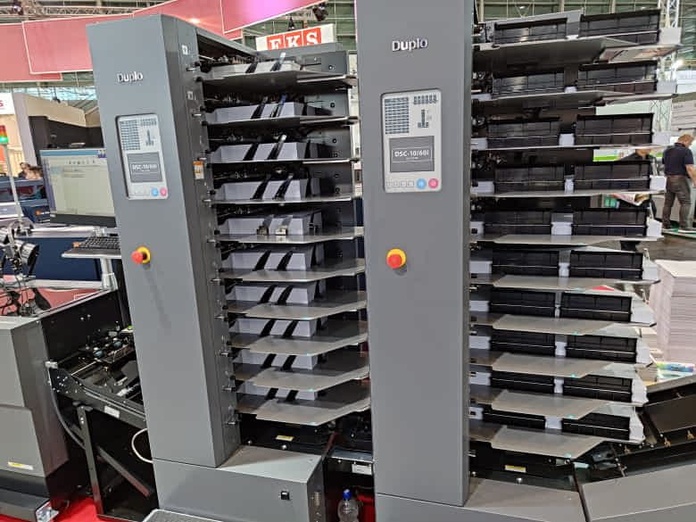 これは製本機システムの構成製品である丁合機です。丁合機には給紙台が10棚配置されており、各給紙台に用紙を積載する必要があります。これを人が行うのは大変な作業となります。協働ロボットで積載できると大きな製造コスト削減となる。